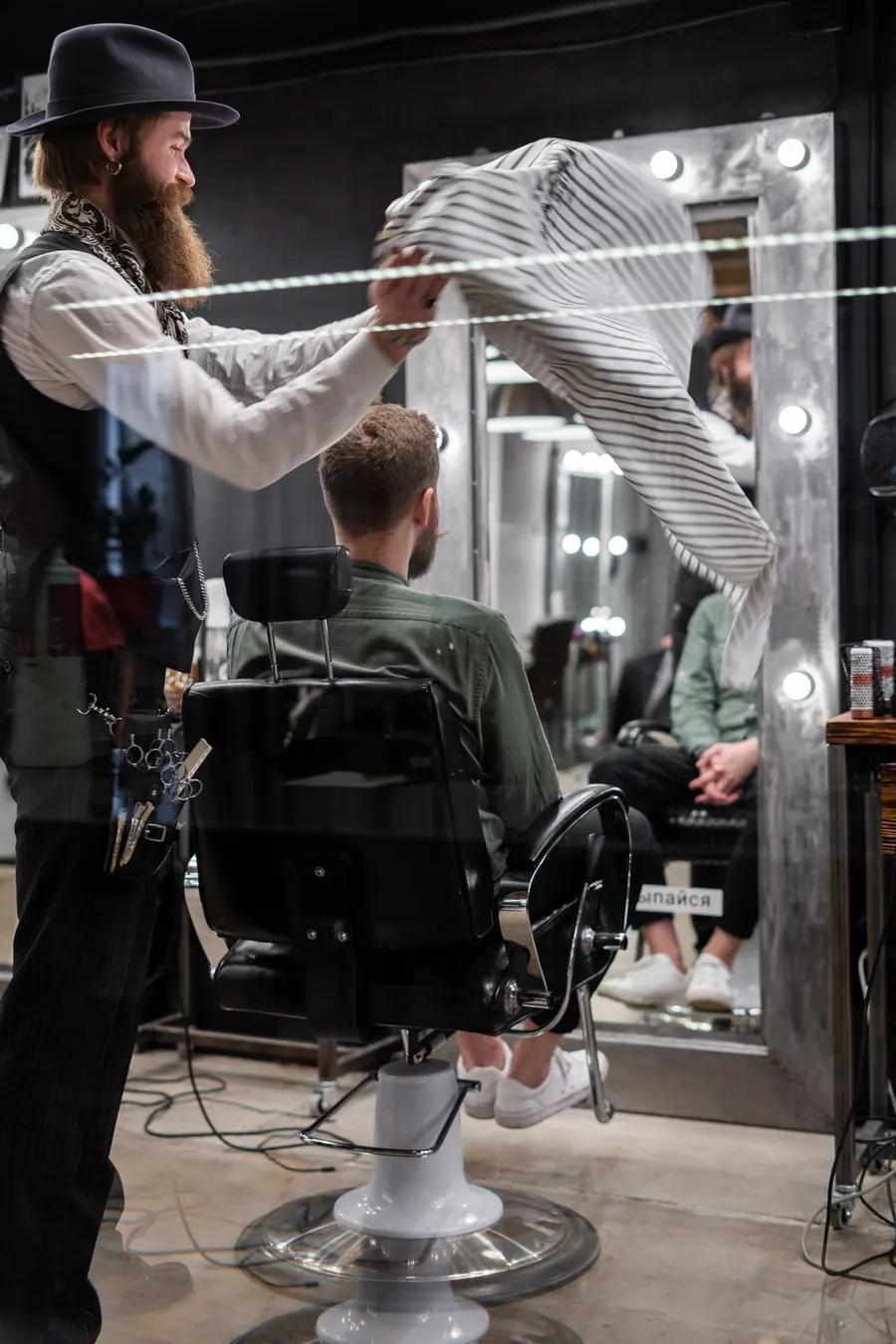 Barber shaking out a striped cape beside a client in a barbershop chair